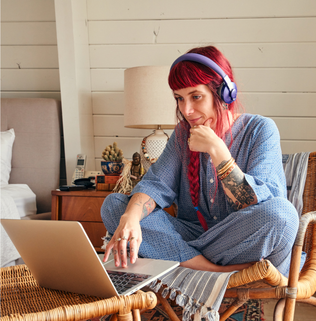 Photo of a woman wearing a headset seated cross-legged on a rattan chair in a bedroom, working on a laptop placed on another chair in front of her