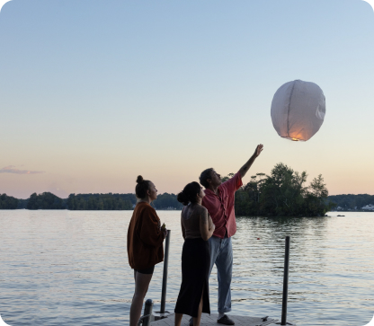 Photo of a trio standing at the edge of a lake at dusk. One of the three has released a dimly glowing lantern to float up into the sky, while the other two observe