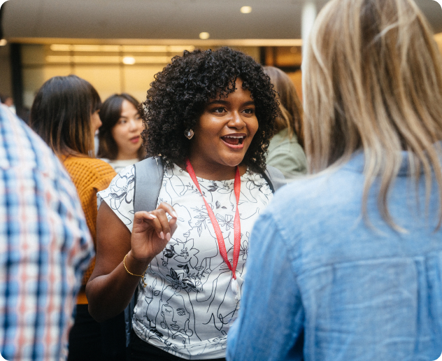 A photo of a woman energetically speaking to another woman in the foreground. The speaker is in focus and surrounded by others also engaged in separate conversations.